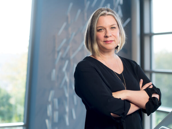White woman standing with arms crossed. She has medium length blonde hair and is dressed in black with a gold necklace. She stands in front of a window and the wall behind her has a reflection of a geometic pattern.