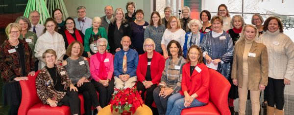 Photo of 31 men and women in clothing of various colors sitting on red couches and standing in the S. Jim and Darla Farha Great Hall