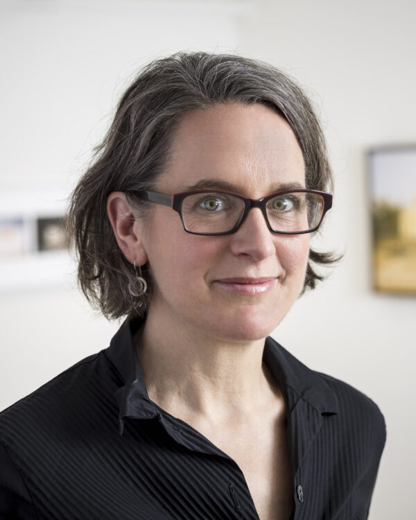 Woman with short dark hair, wearing dark brown-rimmed glasses, dressed in a black, collared, v-neck