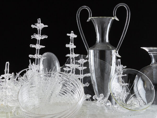 Several dozen clear glass bowls, receptacles and vases assembled on a long white table against a black gallery wall in the background
