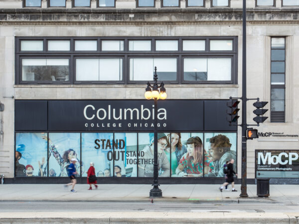 Street scene with stone building facade. Sign in the center over a series of windows identifies building as Columbia College Chicago. Underneath the sign in the windows is a collage of young people and text Stand Out, Stand Together. At the far right at street level is a sign, white on black, MoCP in large letters over Museum of Contemporary Photography in smaller letters. At the far left is two people walking toward to right, In the center are two more people on sidewalk. Center right are two more people walking on the sidewalk and in the right corner is a man standing at the corner facing to cross the street.