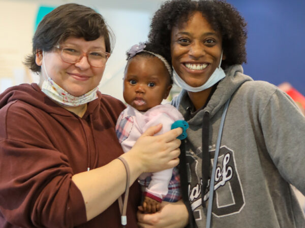Two women, one black wearing a grey sweatshirt, and one white, earing a maroon sweatshirt and wearing glasses, stand and smile for the camera as they hold a small, black girl wearing a pink sweatshirt