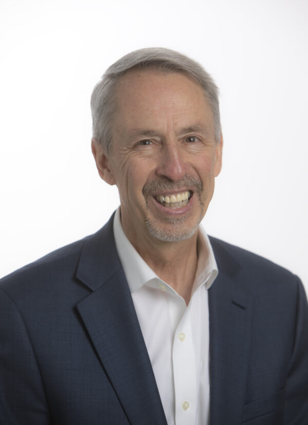 Head and shoulders photo of a man with a big smile, graying hair, wearing a dark suit jacked and a white, open-necked shirt
