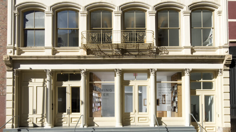 Two stories of a building on white stone. Gray steops span the width of the building on the street level. Five columns are across the first level, with wood and glass doors. The second level has recessed window with carved arches and an metal balcony with round scrolls. Doors in th center ar flanked with tall windows with writing. On the left, the window says The Drawing Center.