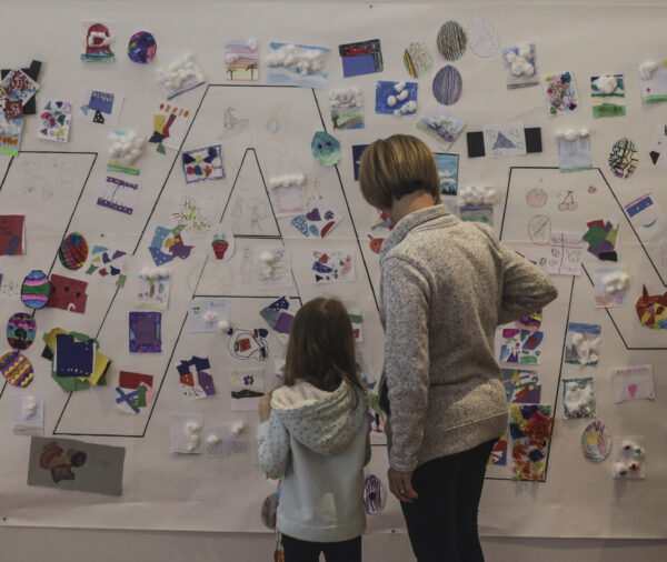 Young girl and woman with backs the the camera, looking at a white wall covered with notes and small pictures